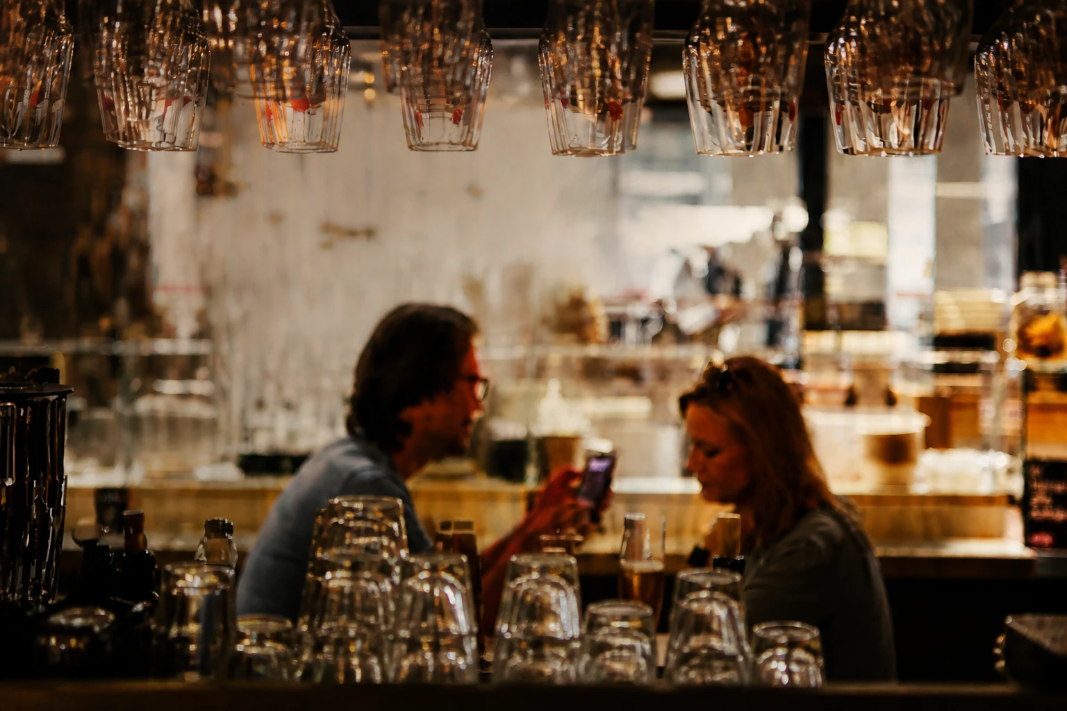 Sfeervol barinterieur in warm avondlicht, met hangende glazen boven de toog en gasten aan tafel